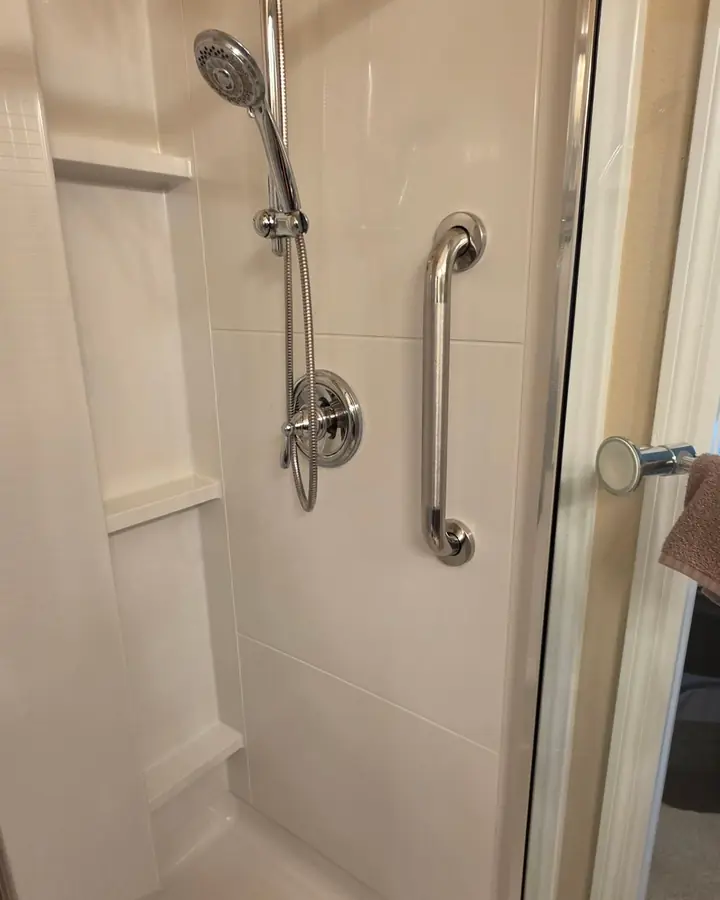 Vertical stainless grab bar installed on a white fiberglass shower surround next to a handheld shower