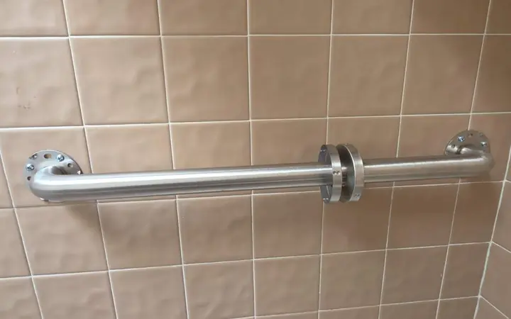Long stainless grab bar on textured tile with the flange covers off, showing the screws that anchor it to the wall
